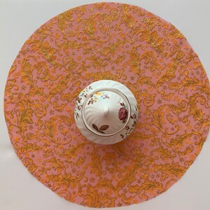 May include: A pink and gold round table runner with a floral pattern. The runner has a white porcelain sugar bowl with a floral design in the center.