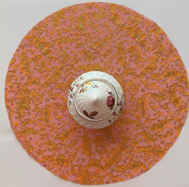 May include: A pink and gold round table runner with a floral pattern. The runner has a white porcelain sugar bowl with a floral design in the center.