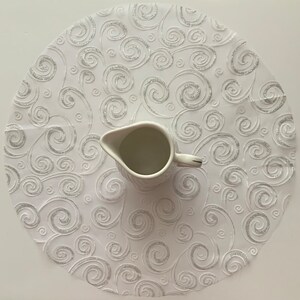 May include: White round table runner with a silver swirl pattern. A white ceramic creamer sits in the center of the runner.