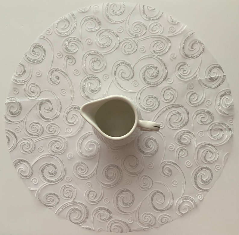 May include: White round table runner with a silver swirl pattern. A white ceramic creamer sits in the center of the runner.