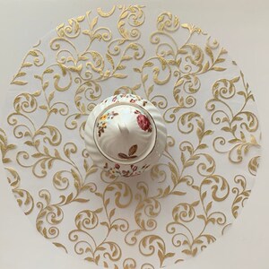 May include: A white ceramic sugar bowl with a floral design sits on a white and gold round paper doily with a swirling floral pattern.