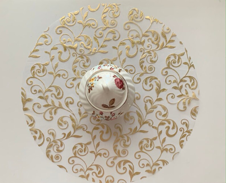 May include: A white ceramic sugar bowl with a floral design sits on a white and gold round paper doily with a swirling floral pattern.