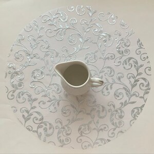 May include: A white lace tablecloth with a silver floral pattern. A white ceramic creamer sits on top of the tablecloth.