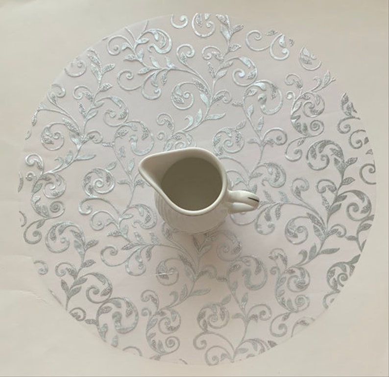 May include: A white lace tablecloth with a silver floral pattern. A white ceramic creamer sits on top of the tablecloth.