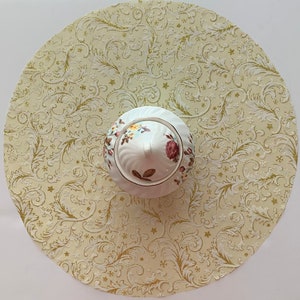 May include: A white ceramic sugar bowl with a floral design and gold accents sits on a cream-colored tablecloth with a gold and white floral pattern.