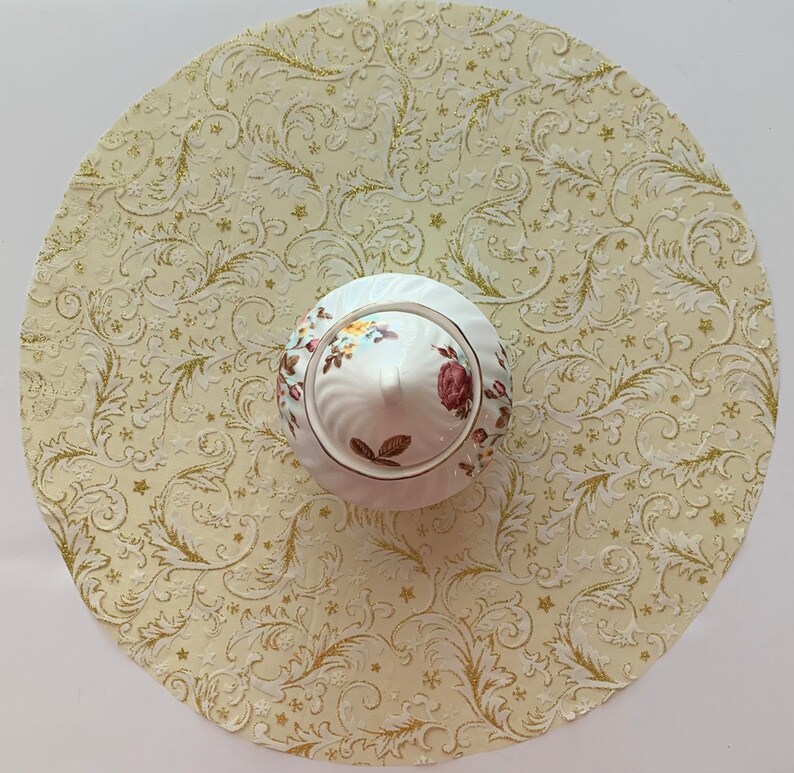 May include: A white ceramic sugar bowl with a floral design and gold accents sits on a cream-colored tablecloth with a gold and white floral pattern.