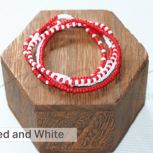 May include: Red and white beaded bracelet on a wooden stand. The bracelet is made of small, round beads in a repeating pattern of red and white.