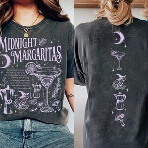 May include: A charcoal gray short-sleeve t-shirt featuring light purple graphic prints. The front displays the text Midnight Margaritas with illustrations of a margarita glass, a frog, a mushroom, and a coffee maker. The back shows a vertical design.