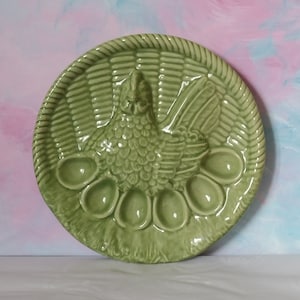 May include: A green ceramic egg plate with a hen and basket design. The plate features a detailed relief of a hen in the centre, surrounded by egg-shaped indentations. The rim has a woven basket pattern. A decorative kitchenware item.