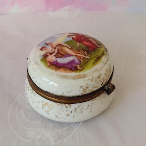 May include: A round white porcelain trinket box with a hinged brass rim. The lid features a colourful painted scene of three figures in a landscape. The exterior of the box is decorated with delicate gold filigree patterns around the base and lid.