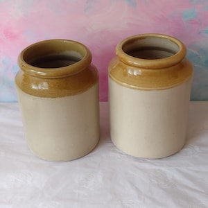 May include: Two cylindrical ceramic storage jars with a two-tone design. Each jar features a smooth, cream-coloured base and a contrasting tan-coloured rim and neck. The jars have a wide, open top with a thick, rounded lip and a sturdy, flat base.