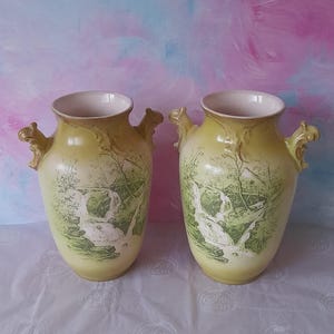 May include: Two antique yellow vases with a landscape design featuring a waterfall and trees. Each vase has a cream-coloured interior and decorative handles. The vases are displayed on a white surface.