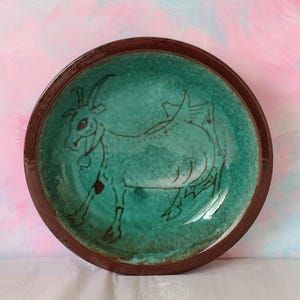May include: A round ceramic bowl with a dark brown rim and a vibrant turquoise interior. The centre of the bowl features a dark brown line drawing of a goat with horns and a bell around its neck, set against a mottled, textured green glaze.