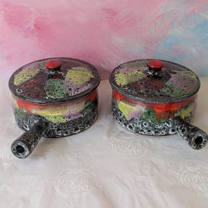 May include: Two round ceramic pots with lids and single handles, featuring a textured black base with a mottled glaze of red, yellow, green, and purple. Each lid has a small red knob, and the overall finish is glossy with a cratered, volcanic texture.