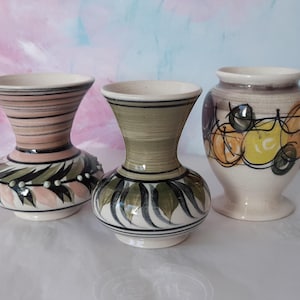 May include: Three ceramic vases with distinct designs. The left vase has pink horizontal stripes and green leaf patterns. The centre vase is olive green with dark leaf motifs. The right vase features yellow and black circular abstract patterns.