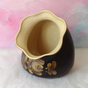 May include: A dark brown ceramic vase with a cream interior and a scalloped rim. The vase has hand-painted gold floral designs. The vase is a decorative item.