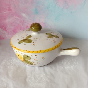 May include: White ceramic soup tureen with a lid and handle, featuring hand-painted floral designs in green and brown. The rim has a yellow and orange pattern. The lid has an olive-green knob. A decorative kitchenware item.