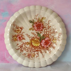 May include: A round, cream-coloured ceramic plate with a scalloped edge and a gold-trimmed rim. The centre features a detailed floral pattern with pink, yellow, and orange flowers, green leaves, and brown lattice-style decorative scrollwork.