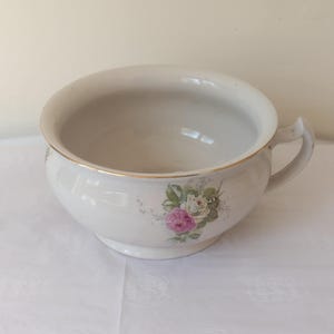 May include: A white ceramic chamber pot featuring a floral decal of pink and white roses with green leaves on the front. The bowl has a rounded handle on the right side and a thin gold-coloured trim along the top rim of the basin.