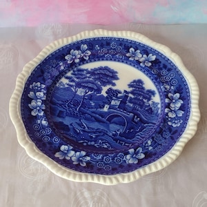 May include: A round ceramic plate with a scalloped, cream-coloured rim. The centre features a detailed blue and white transferware landscape scene depicting a stone bridge, trees, and buildings. The border is decorated with blue floral patterns.