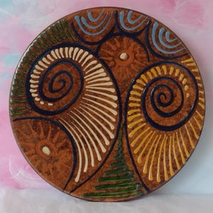 May include: A round ceramic plate with a brown mottled glaze featuring hand-painted designs. The surface displays two large dark blue spirals, radiating lines in cream and yellow, a green triangular pattern, and sun-like motifs in light blue and brown.
