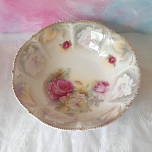 May include: A round porcelain bowl with a scalloped, gold-trimmed edge. The interior features a hand-painted floral design with large pink and cream roses, smaller red rosebuds, and soft green leaves against a cream-coloured background.