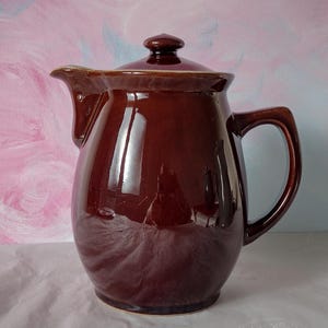 May include: A glossy dark brown ceramic jug with a rounded body, a curved handle, and a matching lid. The jug features a prominent spout and a smooth, reflective finish that catches the light, showcasing its solid, deep brown colour throughout.