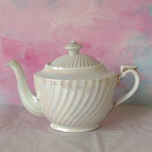 May include: A white, iridescent teapot with a lid, spout, and handle. The teapot features a spiral pattern and gold detailing. The background is a soft pink and blue watercolour wash.