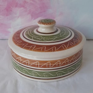 May include: A round ceramic lidded container. The lid and body are adorned with bands of brown, green, and white. The lid features a small knob. The container, with a cream base, is likely for storage.
