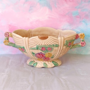 May include: A beige ceramic planter with floral designs in pink, yellow, and green. The planter has two handles and a scalloped rim. The base is also decorated with green leaves. The background is a watercolour wash of pink and blue.