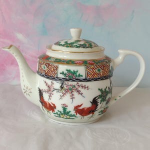 May include: A white porcelain teapot featuring a hand-painted design of two red roosters, pink flowering branches, and green foliage. The upper rim is decorated with a geometric pattern in orange, black, and gold, topped with a matching lid.