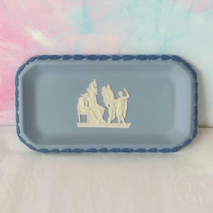 May include: A rectangular, pale blue decorative tray with a raised border and a white bas-relief scene of classical figures. The tray's border has a repeating leaf design. The background is a soft, pastel colour.