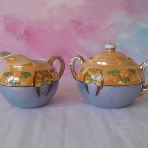 May include: A vintage ceramic creamer and sugar bowl set. The set features a light blue base with an orange top, adorned with owl and palm tree designs. The handles and rims are accented with gold trim, creating an elegant aesthetic.