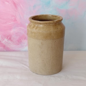 May include: A cylindrical stoneware crock featuring a smooth, tan-coloured rim and a vertically ribbed body in a lighter beige tone. The vessel has a wide, open top and a sturdy base, showcasing a two-toned finish with a distinct texture on the exterior.