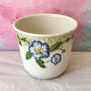May include: A cream-coloured ceramic planter featuring a raised, embossed lattice pattern around the upper rim. The exterior is decorated with hand-painted blue flowers with yellow centres and green leaves, set against a smooth, glossy finish.