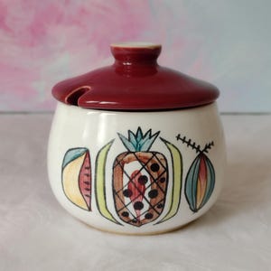 May include: A white ceramic jar with a maroon lid and fruit illustrations. The jar features a pineapple, watermelon, and other fruit designs in black outline with colourful accents. The lid has a small handle and a slot for a spoon.