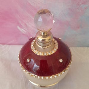 May include: An ornate perfume bottle with a clear crystal stopper. The bottle is burgundy, cream, and gold, with a ring of clear rhinestones. The round bottle has a decorative design. The bottle is a vintage style.