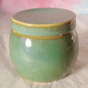 May include: A small, round ceramic jar with a lid. The jar is a glossy, seafoam green colour with a yellow-gold rim. The lid is a darker, muted green. The jar sits on a white, textured surface.