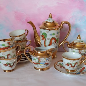 May include: A vintage porcelain tea set with a white base, gold trim, and dragon motifs. The set includes a teapot, sugar bowl, creamer, cups, and saucers. The dragon design features red, green, and blue accents. The set is displayed on a white surface.