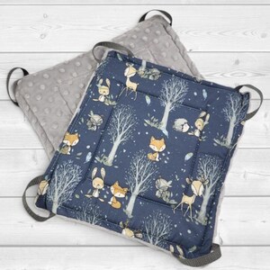 May include: A square seat cushion featuring a dark blue fabric top with a repeating pattern of foxes, deer, and trees in white and orange. The underside is a solid gray fabric with a raised dot texture. Four gray fabric loops are attached to corners.