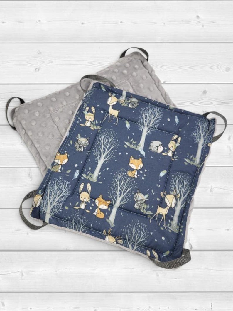 May include: A square seat cushion featuring a dark blue fabric top with a repeating pattern of foxes, deer, and trees in white and orange. The underside is a solid gray fabric with a raised dot texture. Four gray fabric loops are attached to corners.