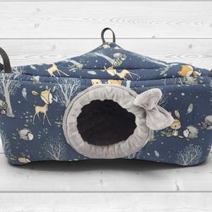 May include: A dark blue quilted pet hammock featuring a forest-themed print with deer, hedgehogs, and trees. The hammock has a circular entrance lined with soft gray fabric, a small gray fabric bow detail, and three black nylon hanging straps.