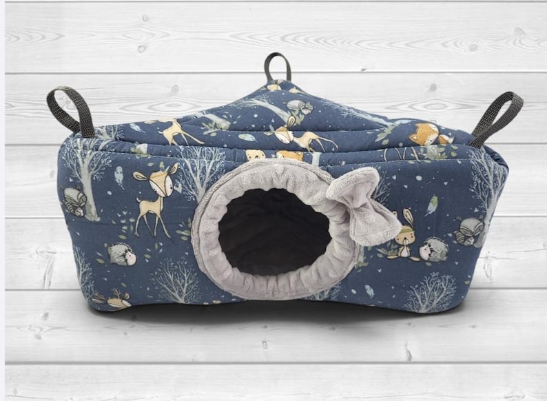 May include: A dark blue quilted pet hammock featuring a forest-themed print with deer, hedgehogs, and trees. The hammock has a circular entrance lined with soft gray fabric, a small gray fabric bow detail, and three black nylon hanging straps.