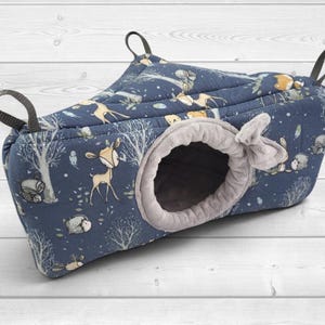 May include: A triangular hanging pet hammock featuring a dark blue fabric with a printed pattern of deer, hedgehogs, and trees. The entrance is circular with a soft light gray plush lining and a small fabric bow detail. Three black straps are attached.