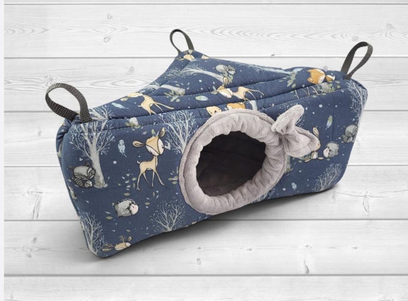 May include: A triangular hanging pet hammock featuring a dark blue fabric with a printed pattern of deer, hedgehogs, and trees. The entrance is circular with a soft light gray plush lining and a small fabric bow detail. Three black straps are attached.