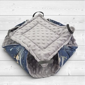 May include: A square-shaped pet bed featuring a dark blue exterior fabric with a printed pattern of trees and animals. The interior is lined with a soft, light gray minky fabric with a raised dot texture. Four black fabric loops are attached.