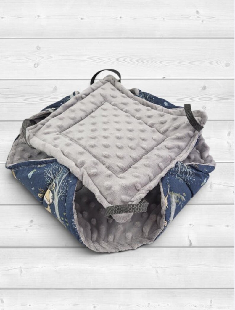 May include: A square-shaped pet bed featuring a dark blue exterior fabric with a printed pattern of trees and animals. The interior is lined with a soft, light gray minky fabric with a raised dot texture. Four black fabric loops are attached.