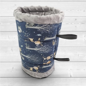 May include: A cylindrical fabric sleeve featuring a dark blue exterior with a repeating pattern of stylized deer and trees in light brown and white. The top and bottom edges are trimmed with a soft, textured grey fabric and two black straps.