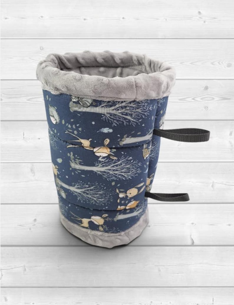 May include: A cylindrical fabric sleeve featuring a dark blue exterior with a repeating pattern of stylized deer and trees in light brown and white. The top and bottom edges are trimmed with a soft, textured grey fabric and two black straps.