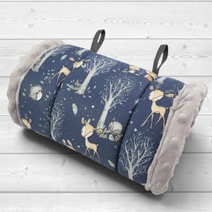 May include: A rolled-up fabric mat featuring a dark blue background with a repeating pattern of light brown deer, grey trees, and small animals. The ends of the mat are finished with a textured, light grey plush fabric and two black fabric loops.